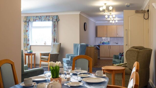 <p>Wide shot of an empty communal living room in a residential care home in North East England. The room is tidy, featuring a dining table set with crockery, several armchairs and a small kitchen area at the far end. There is a window which illuminates the space with natural light. Videos similar to this scenario available.</p>